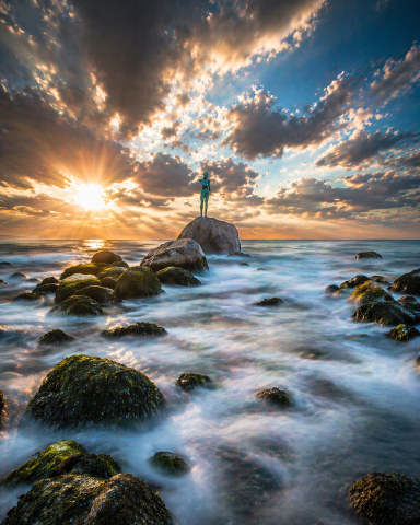 Statue am Selliner Strand Eine Statue steht auf einem Felsen im Meer, umgeben von Wellen und Sonnenlicht.