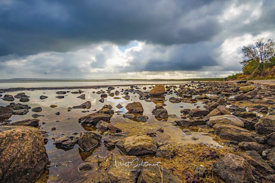 Wilde Ostseeküste auf Rügen mit beeindruckendem Wolkenhimmel Schmale Küste mit Steinen und Wolken am Himmel, reflektiert im Wasser.